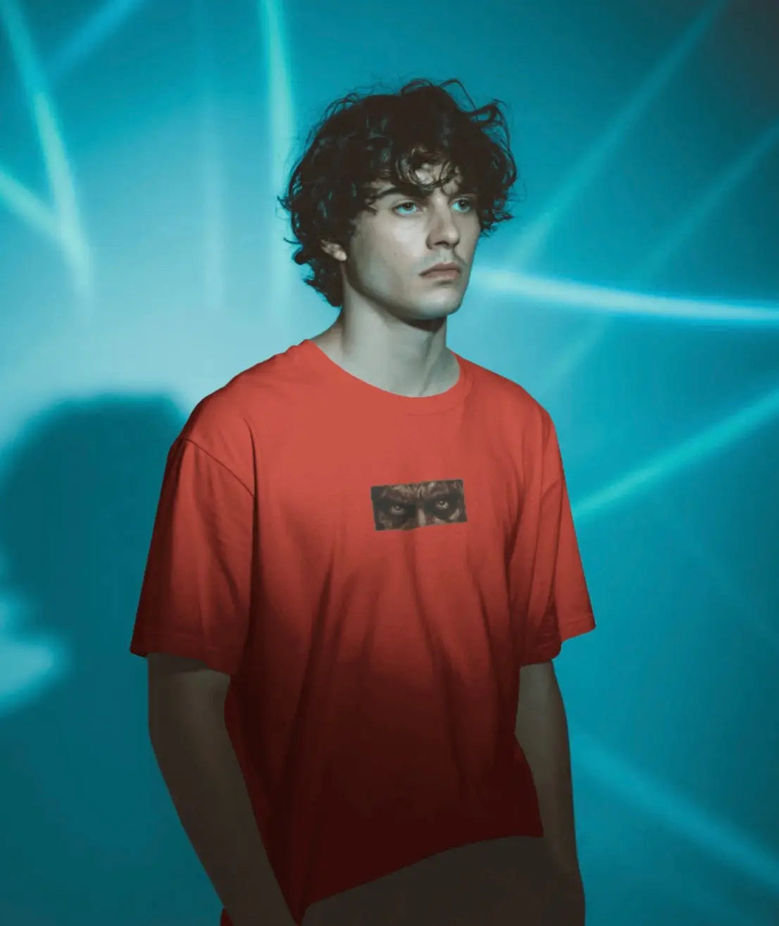 “A young man with dark curly hair models a red t-shirt against a blue background with light projections. The shirt features a small rectangular graphic on the chest showing a pair of intense, staring eyes.”